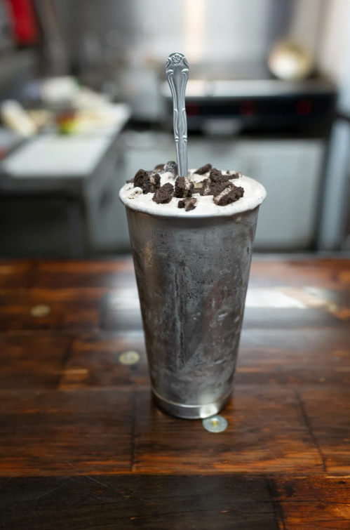 A tall metal cup filled with a creamy milkshake topped with crushed cookies and a silver spoon.
