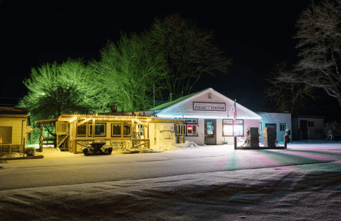 A snow-covered gas station at night, illuminated by colorful lights and surrounded by trees.