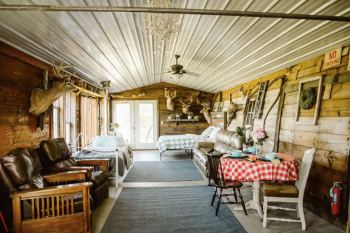 Cozy rustic interior with wooden walls, a checkered table, sofas, and decorative elements like antlers and a chandelier.