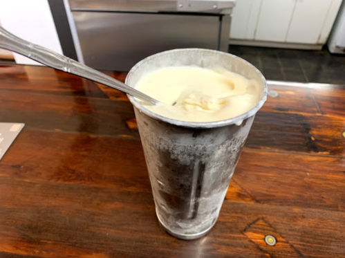 A metal cup filled with creamy dessert, resting on a wooden table with a spoon inside.