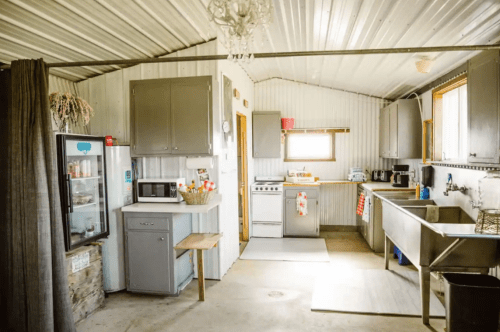 A rustic kitchen with gray cabinets, a fridge, microwave, and stainless steel sinks, featuring a cozy, inviting atmosphere.