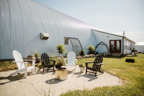 A cozy outdoor seating area with black and white chairs around a fire pit, next to a large metal building.