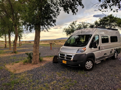 A silver camper van parked among trees at a scenic campsite with a cloudy sky in the background.