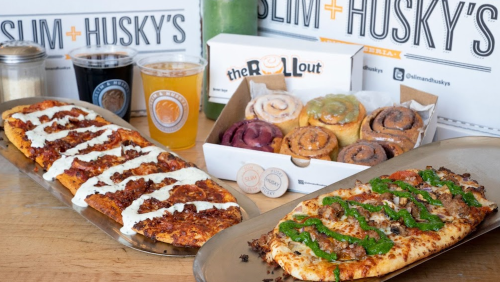 Two pizzas on metal trays, one topped with meat and drizzled with sauce, alongside a box of colorful cinnamon rolls and drinks.