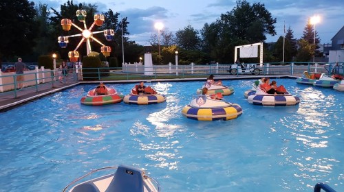 A group of people in colorful bumper boats on a pool, with a ferris wheel and evening sky in the background.