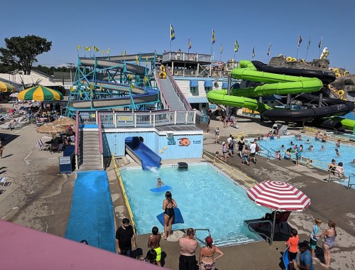 A busy water park with slides, pools, and people enjoying the sun under colorful umbrellas.