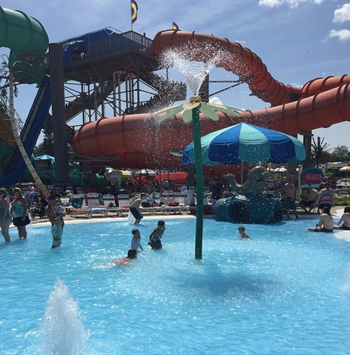 A water park scene with a splash pad, water slides, and people enjoying the pool on a sunny day.
