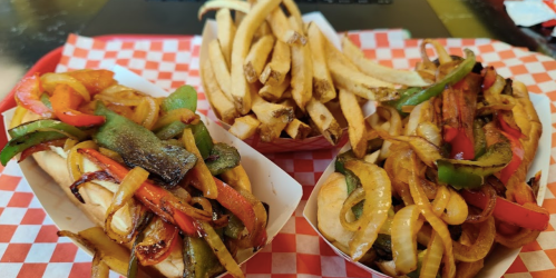 Three loaded hot dogs topped with grilled peppers and onions, served with a side of golden French fries on a red checkered tray.