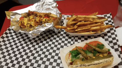A plate featuring a loaded hot dog in foil, a hot dog with toppings, and a side of crispy French fries on checkered paper.