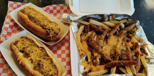 Two loaded chili cheese hot dogs and a tray of cheese-covered fries in a takeout container on a checkered paper.