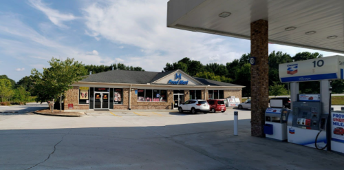 A convenience store with a gas station, surrounded by trees and a clear blue sky. Cars are parked in front.