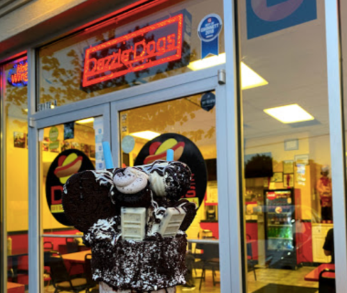 A colorful ice cream cone topped with cookies and sprinkles in front of a dessert shop with a neon sign.