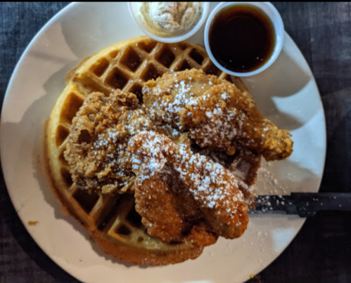 A plate featuring a golden waffle topped with crispy fried chicken, powdered sugar, and sides of syrup and butter.