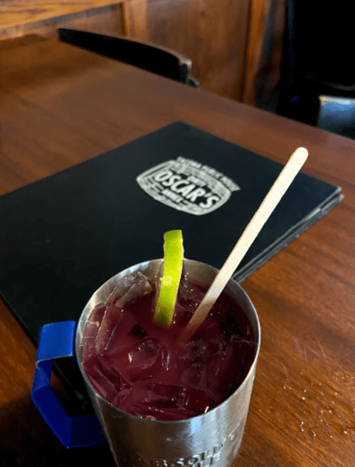 A cocktail with ice and a lime wedge in a metal cup, placed on a wooden table next to a menu.