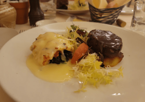 A plate featuring a portion of steak with sauce, accompanied by vegetables and a cheesy dish on the side.