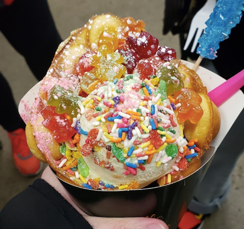 A colorful dessert with ice cream, sprinkles, gummy candies, and a waffle cone, held in a hand.
