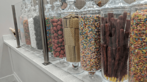 A row of glass jars filled with colorful candies, sprinkles, and snacks on a countertop.