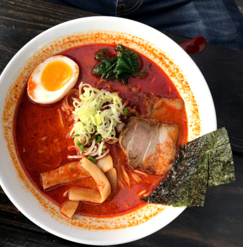 A bowl of spicy ramen topped with sliced pork, a soft-boiled egg, green onions, and seaweed.