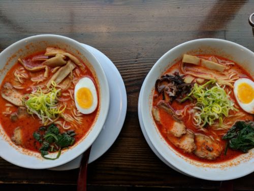 Two bowls of ramen with red broth, noodles, sliced egg, vegetables, and meat, served on a wooden table.