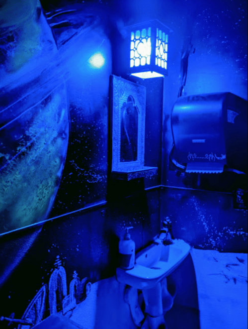 A blue-lit bathroom with a cosmic mural, featuring a sink, mirror, and paper towel dispenser.