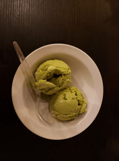 A white bowl containing two scoops of green ice cream with a clear plastic spoon on a dark wooden surface.