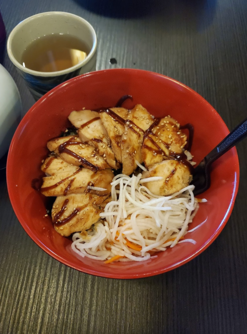 A red bowl filled with sliced grilled chicken, rice noodles, and drizzled sauce, accompanied by a cup of tea.