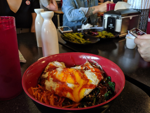 A bowl of bibimbap topped with an egg and sauce, with drinks and edamame in the background at a restaurant.