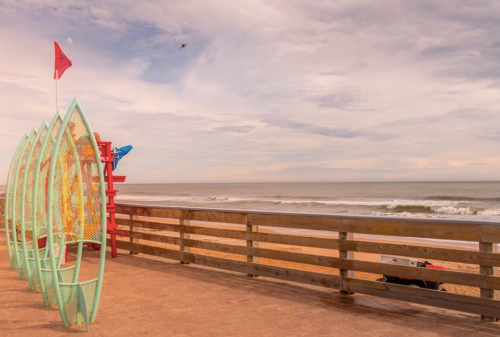 Colorful surfboards stand on a wooden deck overlooking a calm beach and ocean under a cloudy sky.