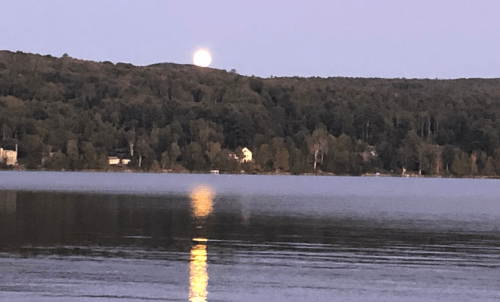 A serene lake at dusk, with a full moon rising over a tree-lined hill, reflecting on the water's surface.