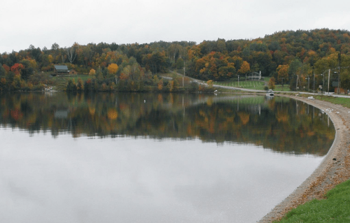 A calm lake reflects colorful autumn trees and a shoreline with a gravel path, under a cloudy sky.