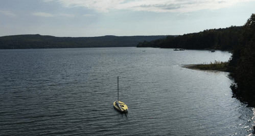A calm lake scene with a single sailboat and distant hills under a partly cloudy sky.