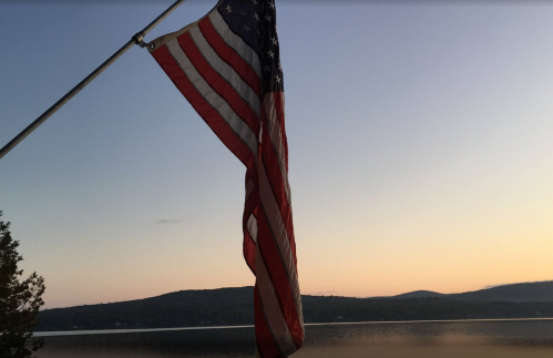 A U.S. flag waves in the foreground with a serene lake and mountains at sunset in the background.