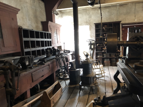 A vintage workshop filled with tools, machinery, and wooden furniture, illuminated by natural light from a window.