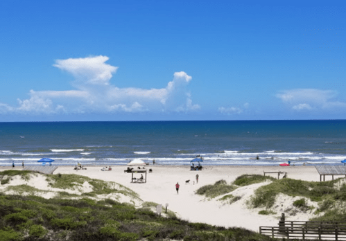 A sunny beach scene with people, umbrellas, and gentle waves under a clear blue sky.