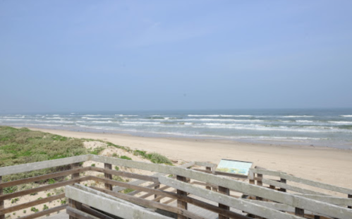 A sandy beach with gentle waves, viewed from a wooden deck under a clear blue sky.