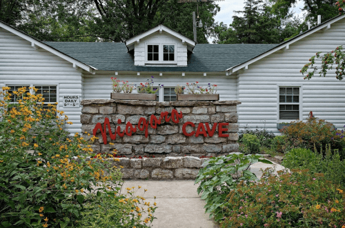 Sign for Niagara Cave in front of a white building, surrounded by colorful flowers and greenery.