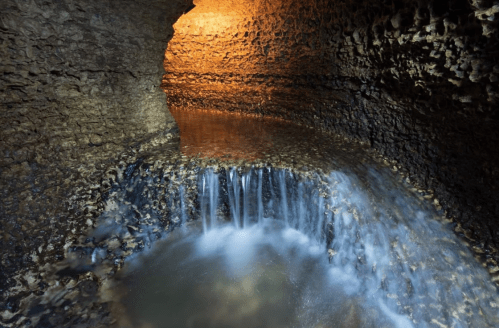 A serene underground cave with a small waterfall flowing into a calm pool, illuminated by warm light.