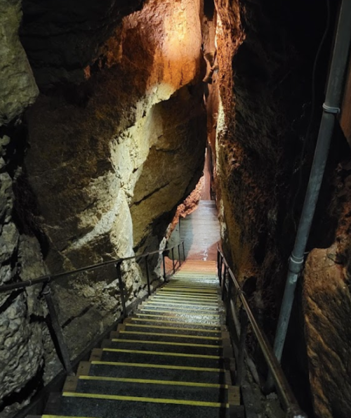 A narrow staircase descends into a rocky cave, illuminated by warm lights along the walls.