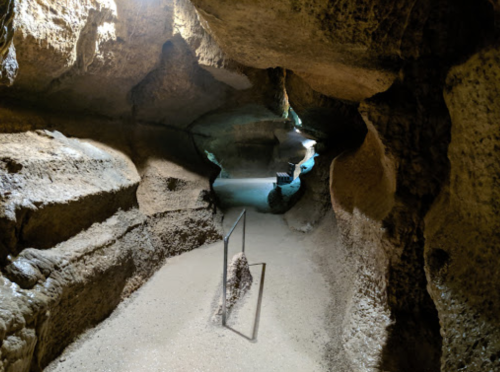 A dimly lit cave interior with rocky walls and a sandy floor, featuring a railing along the path.