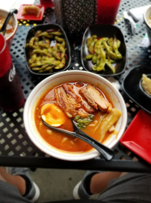 A bowl of ramen with pork, egg, and vegetables, surrounded by edamame and drinks on a table.
