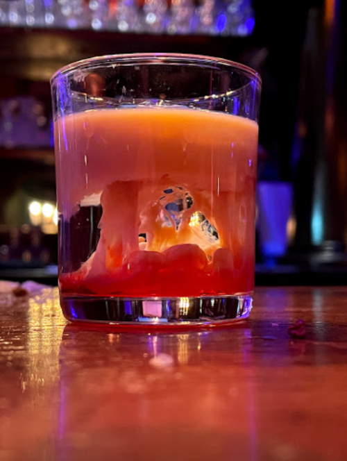 A glass of layered drink with orange and red hues, sitting on a bar counter with a blurred background.