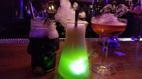 Three unique cocktails on a bar: a skull mug with dark drink, a glowing green drink in a flask, and a classic cocktail.
