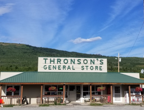 A general store with a green roof and flower boxes, set against a backdrop of mountains and a blue sky.