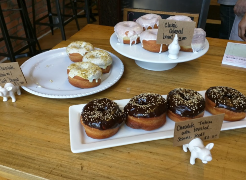 A display of assorted donuts: chocolate tahini, classic glazed, and a frosted variety, with price tags on a wooden table.
