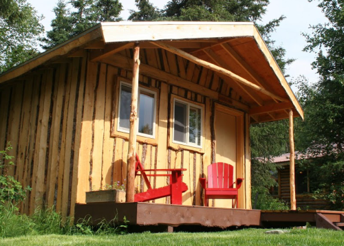 A rustic wooden cabin with a porch, featuring red chairs and surrounded by trees.