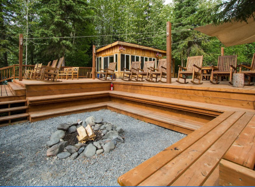 A wooden deck with rocking chairs surrounds a fire pit, set in a forested area with a cabin in the background.