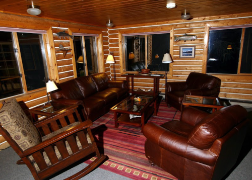 Cozy log cabin living room with leather sofas, a rocking chair, and large windows, illuminated by soft lamps.