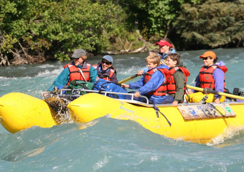 A group of six people in life jackets enjoying a rafting adventure on a river, surrounded by lush greenery.