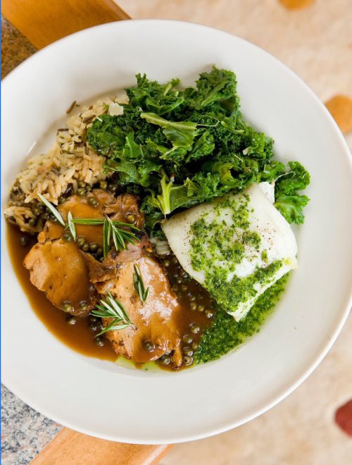 A bowl featuring rice, kale, and tender meat in a savory sauce, garnished with herbs and a green sauce.