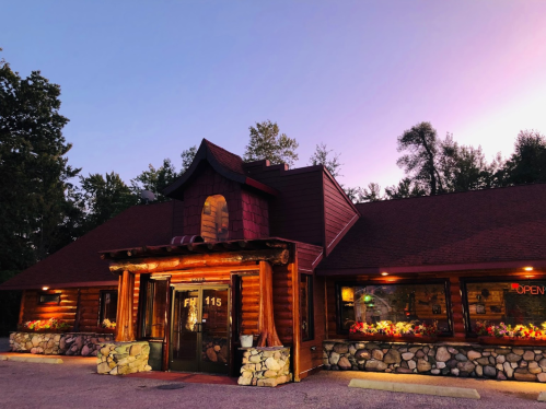 A cozy log cabin restaurant with a welcoming entrance, surrounded by trees and illuminated by evening light.
